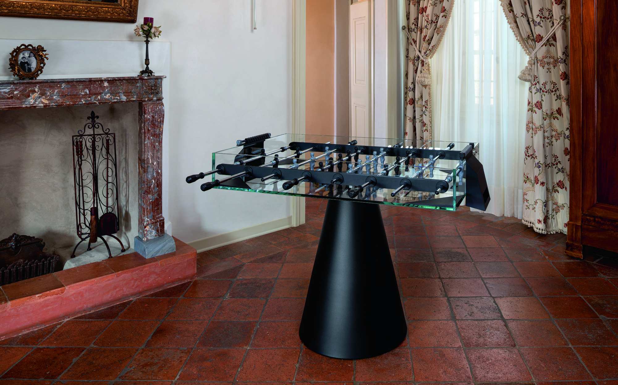 FAS Pendezza Ghost glass foosball table with black cone base beside a classic fireplace